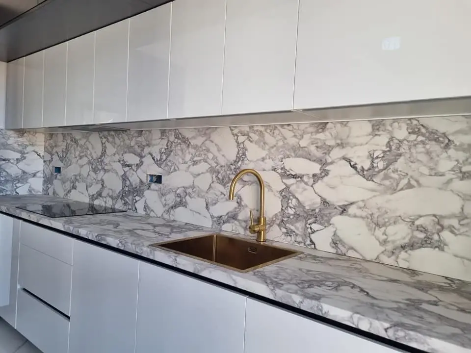 Detalj kuhinje: bijele fronte bez ručki, zidna obloga s uzorkom mramora i zlatna slavina